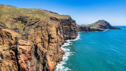 Fototapeta na wymiar A steep, rocky coastline with light blue ocean water at its base. Visible are green grassy areas and paths along the cliffs. It's the beauty of nature in Madeira, the PR8 trail.
