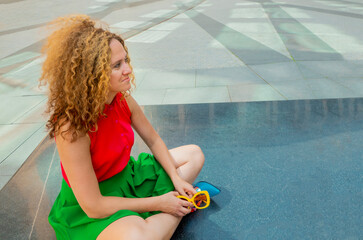 Fashionable woman in bright clothes, with long blond curly hair, is sitting in the lotus position on the street basking in the sun and resting against the background of a modern business center.