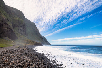 Rocky coastline of Madeira with green slopes, white beach, and undulating sea under a white sky with clouds. It's a landscape that showcases the pristine beauty of nature.