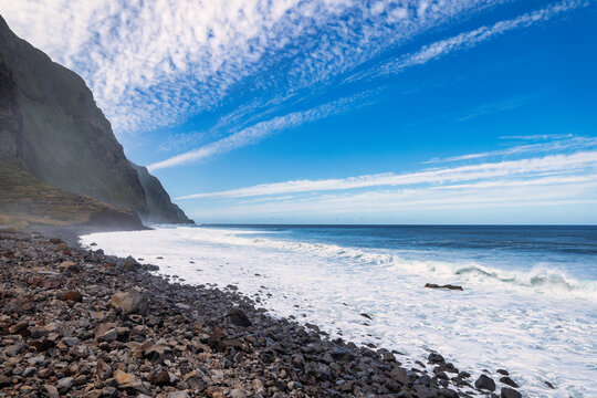 The rocky coast of Madeira, as seen from the Achadas da Cruz viewpoint. Green slopes, a white beach, and blue sea are visible under a partly cloudy sky. The overall mood of the picture is calm and