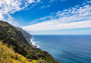 The photo depicts a steep, green mountainside in Madeira, sloping down to the blue sea. Waves crash against the rocky coast, and dense clouds dominate the scene, creating an image of untouched nature.