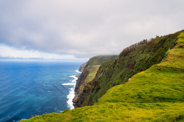 The photo depicts a steep, green mountainside in Madeira, sloping down to the blue sea. Waves crash against the rocky coast, and dense clouds dominate the scene, creating an image of untouched nature.