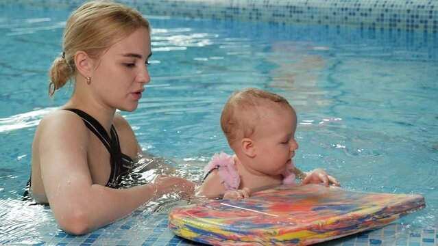 Close-up of a little one-year-old girl with her mother in the pool at a swimming lesson for newborns. The girl is holding onto a foam pad for swimming.