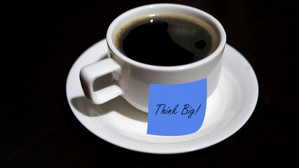 Coffee with text message - Think Big. Closeup of a white cup of black coffee on plate and motivational quote on a blue sticky paper note on dark table background. Monday coffee concept.