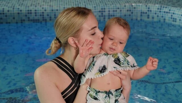 A young caring mother holds her one-year-old daughter in her arms and kisses her in the pool. The mother takes care of the child's health.