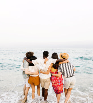 Rear view of young friends hugging each other in front of the sea on summer vacation. Holiday lifestyle concept.