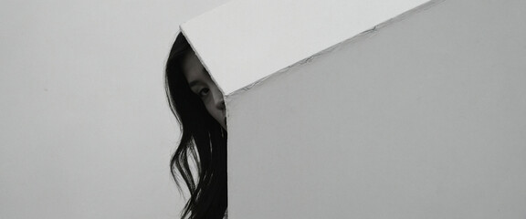 A woman with long, dark hair is peeking from behind a white wall.