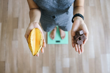 Unrecognized Woman Weighing Body While Holding Star Fruit and Chocolate on Hand. Healthy Diet...