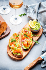 Avocado sandwich with salmon on rye bread with guacamole sauce, arugula and sesame seeds on wooden board, rose wine glass on gray table background, top view