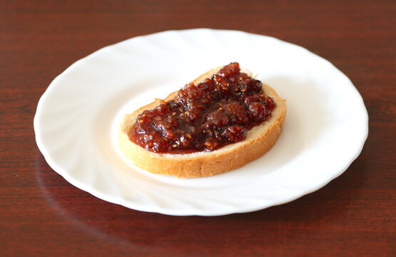 A piece of white bread with raspberry jam is on a white plate