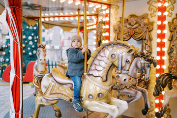 Little girl rides a toy horse on a carousel in the square near a decorated Christmas tree