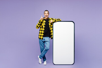 Full body young middle eastern man wears yellow shirt casual clothes big huge blank screen mobile cell phone smartphone with workspace area point index finger up isolated on plain purple background.