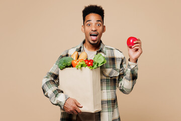 Young shocked happy man wear grey shirt hold apple, paper bag for takeaway mock up with food products isolated on plain pastel light beige background studio. Delivery service from shop or restaurant.