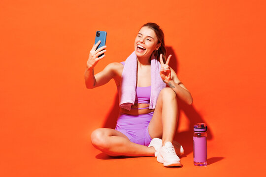 Full body young fitness trainer woman sportsman wear top shorts purple clothes sit in home gym do selfie shot on mobile cell phone isolated on plain orange background. Workout sport fit abs concept.
