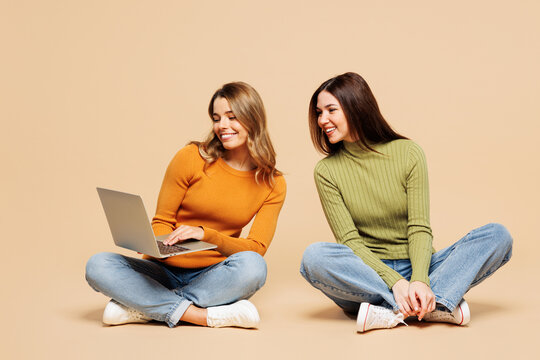 Full body young friends two IT women wear orange green shirt casual clothes together sit hold use work on laptop pc computer isolated on plain pastel light beige background studio. Lifestyle concept.
