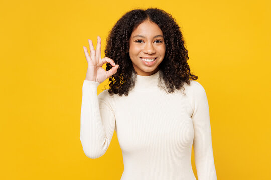 Little smiling happy kid teen girl of African American ethnicity she wear white casual clothes showing okay ok gesture isolated on plain yellow background studio portrait. Childhood lifestyle concept.