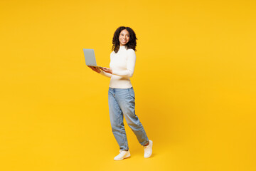 Full body little IT kid teen girl of African American ethnicity in white casual clothes hold use work on laptop pc computer look aside isolated on plain yellow background Childhood lifestyle concept