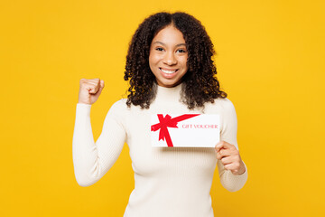 Little kid teen girl of African American ethnicity in white casual clothes hold gift certificate coupon voucher card for store do winner gesture isolated on plain yellow background Childhood concept - obrazy, fototapety, plakaty