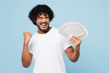 Young happy Indian man he wear white t-shirt casual clothes hold in hand fan of cash money in dollar banknotes do winner gesture isolated on plain pastel light blue cyan background. Lifestyle concept.