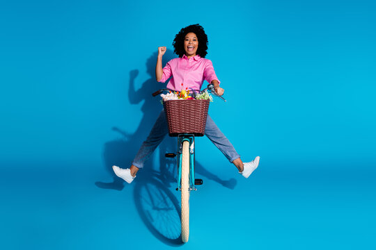 Full length photo of ecstatic nice girl dressed silk shirt jeans riding bike clenching fist have fun isolated on blue color background