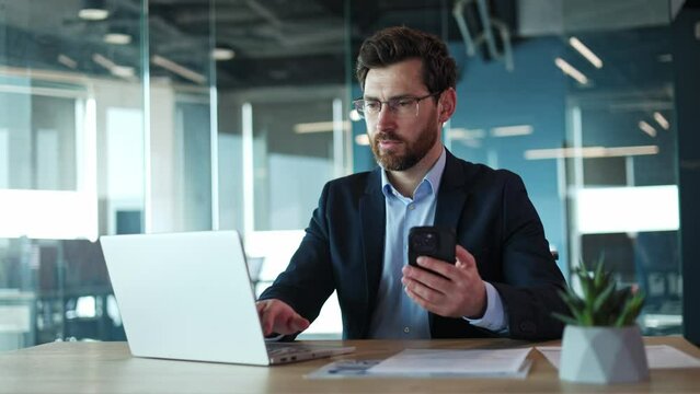 Hardworking male browsing modern smartphone while typing on portable laptop in personal cabinet. Positive caucasian businessman dressed in elegant formal attire and glasses doing job indoors.