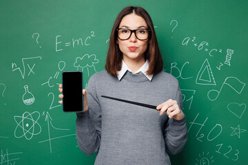 Young smart teacher woman wear grey casual shirt glasses hold use point on blank screen mobile cell phone isolated on green wall chalk blackboard background. Education in high school college concept. - 775740307