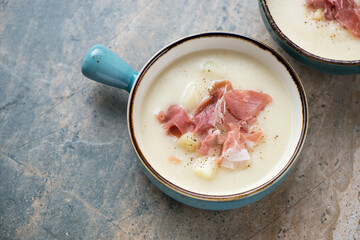 Potato soup with torn prosciutto in a turquoise serving bowl, horizontal shot on a grey and beige granite background, elevated view