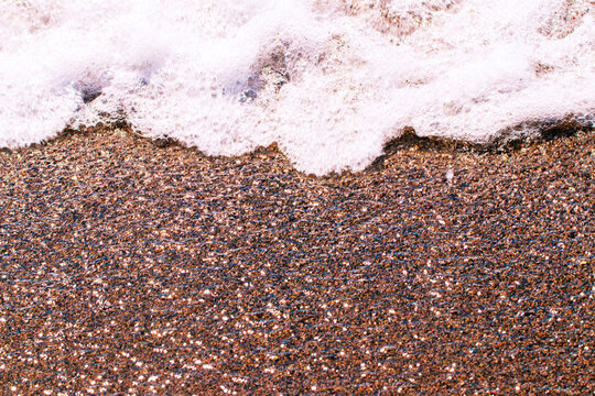 Wave foaming on the sand. Sea waves with white foam on the sandy shore with sun reflections effect. Background texture of mediterranean pabble beach.