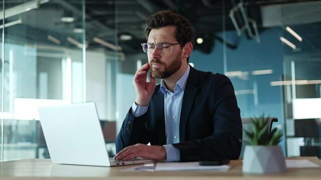 Upset bearded businessman suffering from toothache while sitting at wooden desk on background of glassy wall of corporate office. Disappointed caucasian male in glasses need dental help indoors.