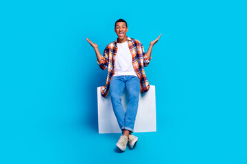 Full length photo of young man sit cube raise hands wear shirt isolated on blue color background