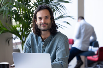 portrait d'un employé de bureau, homme d'affaires sympathique assis qui travaille avec un ordinateur portable. Il regarde la caméra en souriant - 775758334