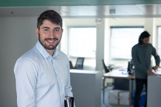 portrait d'un jeune homme employ&eacute; de bureau souriant quoi est assis &agrave; son poste de travail devant son ordinateur. Un coll&egrave;gue est assis derri&egrave;re lui.
