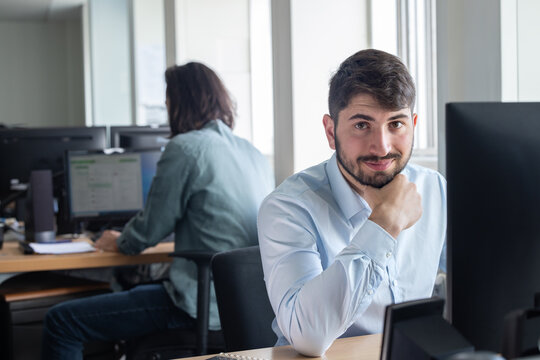 portrait d'un jeune homme employ&eacute; de bureau souriant quoi est assis &agrave; son poste de travail devant son ordinateur. Un coll&egrave;gue est assis derri&egrave;re lui.