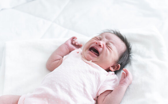 Newborn baby girl crying and screaming, lying on bed at home. Asian Australian infant 19 days old wear clothes, showing unhappy emotion face expression