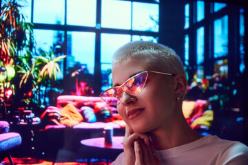 Smiling woman with short hair wearing glasses, with light reflection. Indoor plants backdrop. Watching outdoor holiday celebration. Concept of human emotions