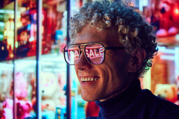 Man with curly hair, wearing glasses reflecting neon 'SALE' sign, smiling. Big shopping sales....