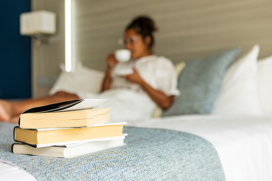 Pile of books on bed, blurred woman drinks coffee at the background