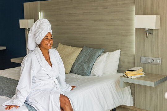 Relaxed middle aged hispanic woman in bathrobe sitting on hotel bed