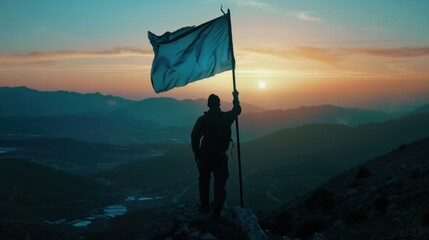Triumphant man hoisting blue flag atop mountain peak at sunset