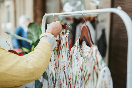 A person's hand is seen perusing through a variety of patterned dresses on a clothing rack at an outdoor boutique stand