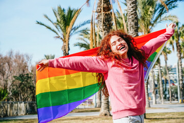 A joyful person with flowing hair spreads their arms wide, the rainbow pride flag a vibrant backdrop to their infectious laughter, amidst a grove of palm trees
