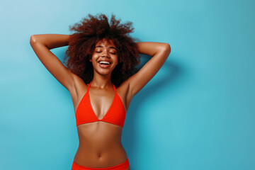 Fototapeta premium Full body portrait of a funny young happy African American woman in red swimsuit going on summer holiday trip and having fun on a blue studio background with copy space. Vacation and travel concept