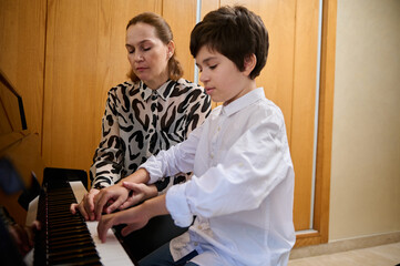 Handsome Hispanic boy in white casual shirt, learning playing piano, sitting at wooden piano forte...