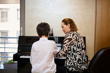Rear view of a music teacher explaining piano lesson to her student. Boy playing piano under the...