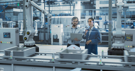 Two Diverse Engineers Standing in a Factory, Using Laptop Computer at an Electronics Factory. Female and Male Specialists Chatting, Monitoring Assembly Robots on a Modern Production Conveyor Belt