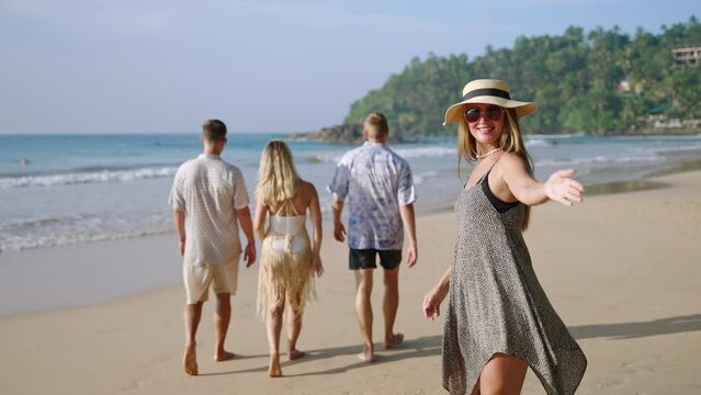 Cheerful caucasian female in sun hat walks by sea on sandy tropical beach with friends group turns, asks to join. Pretty young traveler woman on luxury resort shore smiles cheerfully, invites follow.