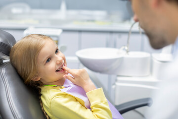Cute little girl showing her milk teeth and waiting for checkup, sitting in chair in dentist office. Early prevention and no fear concept