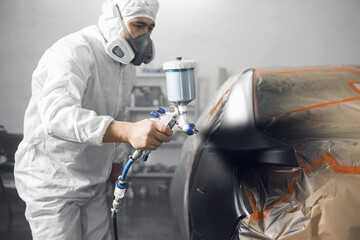Male worker sprays paint with spray gun on car body part in car maintenance service paint room.