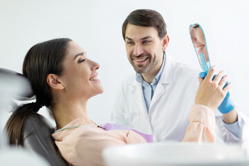 Happy woman patient looking at mirror at her perfect teeth treatment result during dental check up in dentist cabinet, side view
