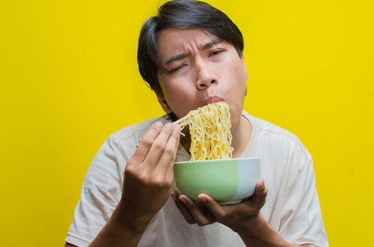 A young Asian man is devouring noodles with his hungry expression turning into one of deep satisfaction.